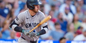 Cody Bellinger, wearing a grey New York Yankees uniform and batting helmet, holds a baseball bat while looking out at the field.