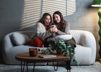 Two women sharing surprising news on a smartphone while sitting on a sofa.