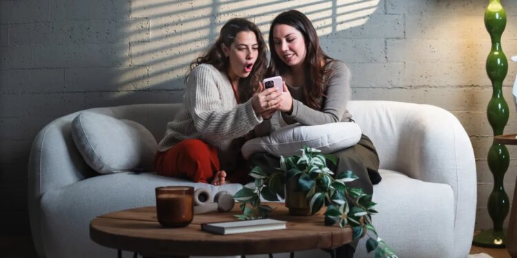 Two women sharing surprising news on a smartphone while sitting on a sofa.