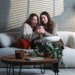 Two women sharing surprising news on a smartphone while sitting on a sofa.