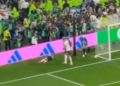 A soccer player lying on the field near the goal with another player standing nearby as fans watch from the stands.