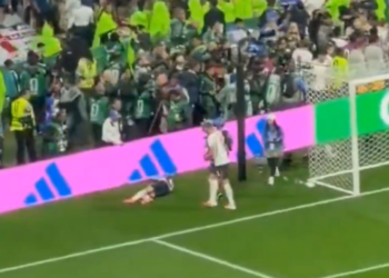 A soccer player lying on the field near the goal with another player standing nearby as fans watch from the stands.