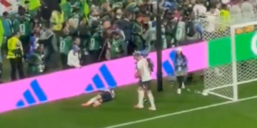 A soccer player lying on the field near the goal with another player standing nearby as fans watch from the stands.