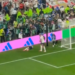 A soccer player lying on the field near the goal with another player standing nearby as fans watch from the stands.