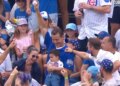 A man in a blue shirt holds a baseball above his head in a crowded baseball stadium, surrounded by smiling fans.