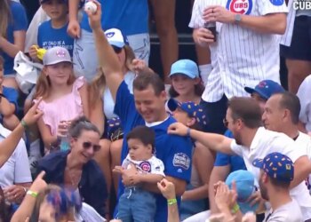 A man in a blue shirt holds a baseball above his head in a crowded baseball stadium, surrounded by smiling fans.