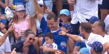A man in a blue shirt holds a baseball above his head in a crowded baseball stadium, surrounded by smiling fans.
