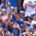 A man in a blue shirt holds a baseball above his head in a crowded baseball stadium, surrounded by smiling fans.
