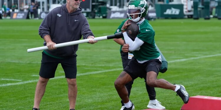 Frank Reich holds a long white pole that Garrett Wilson, in a green helmet and jersey, holds to his stomach with a football.