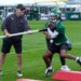 Frank Reich holds a long white pole that Garrett Wilson, in a green helmet and jersey, holds to his stomach with a football.