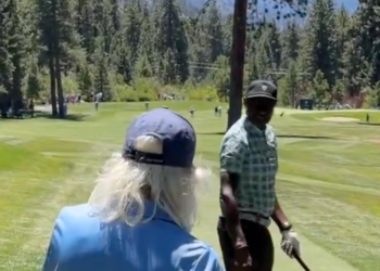 A man with long blonde hair and a blue hat from behind on a golf course.