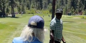 A man with long blonde hair and a blue hat from behind on a golf course.