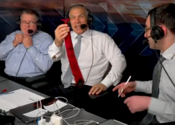 Three men with headsets at a broadcast table, one holding up a red tie.