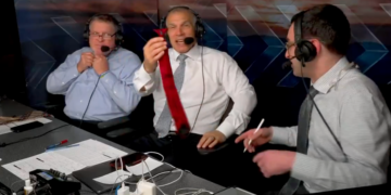 Three men with headsets at a broadcast table, one holding up a red tie.