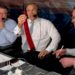 Three men with headsets at a broadcast table, one holding up a red tie.