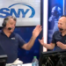 Two men in headphones and microphones, wearing blue SNY shirts, gesturing while broadcasting.