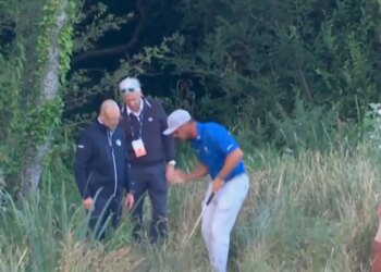 A golfer in a blue shirt with his caddy and other officials in long grass, with a golf leaderboard visible on the right.