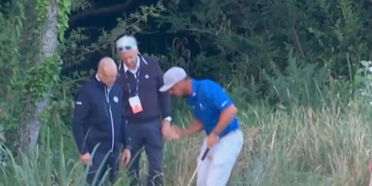 A golfer in a blue shirt with his caddy and other officials in long grass, with a golf leaderboard visible on the right.