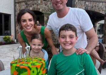 Family of four posing with a birthday cake that reads "BREXTON" in front of a building with a stone archway.