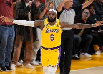 LeBron James in a yellow Lakers jersey with arms outstretched on a basketball court.