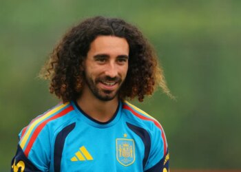 A smiling man with long curly hair and a beard wearing a blue and yellow soccer jersey.