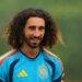 A smiling man with long curly hair and a beard wearing a blue and yellow soccer jersey.