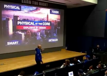 A person in a blue hoodie points at a large screen displaying the words "PHYSICAL AT THE CORE, EXPLOSIVE BY DESIGN" over an image of a city skyline, with students watching.