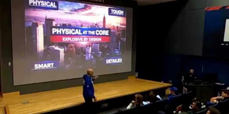 A person in a blue hoodie points at a large screen displaying the words "PHYSICAL AT THE CORE, EXPLOSIVE BY DESIGN" over an image of a city skyline, with students watching.