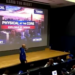 A person in a blue hoodie points at a large screen displaying the words "PHYSICAL AT THE CORE, EXPLOSIVE BY DESIGN" over an image of a city skyline, with students watching.