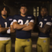 Five male football players in blue jerseys and gold pants stand with their arms crossed.