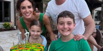 Family of four posing with a birthday cake that reads "BREXTON" in front of a building with a stone archway.