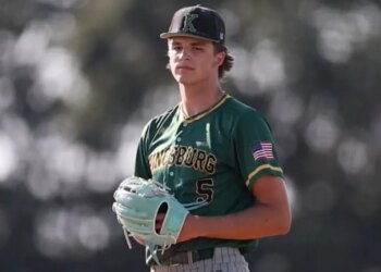 A male baseball player in a green Kingsburg uniform with an American flag patch and a black cap with a green "K" on it, wearing a light blue glove.