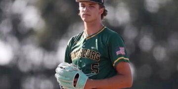 A male baseball player in a green Kingsburg uniform with an American flag patch and a black cap with a green "K" on it, wearing a light blue glove.