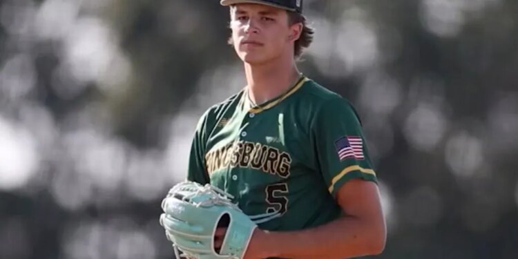 A male baseball player in a green Kingsburg uniform with an American flag patch and a black cap with a green "K" on it, wearing a light blue glove.