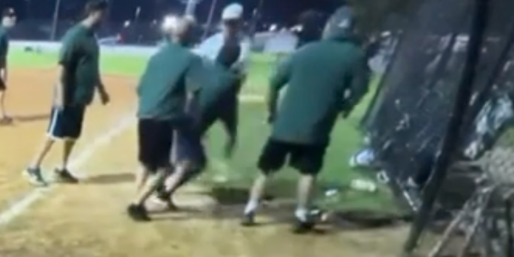 Three men in green shirts and shorts fight another man in a white shirt and dark pants on a baseball field at night.
