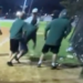 Three men in green shirts and shorts fight another man in a white shirt and dark pants on a baseball field at night.