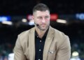 Tim Tebow wearing a tan suit jacket and black shirt with a microphone earpiece.