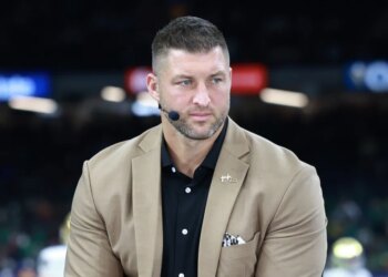 Tim Tebow wearing a tan suit jacket and black shirt with a microphone earpiece.