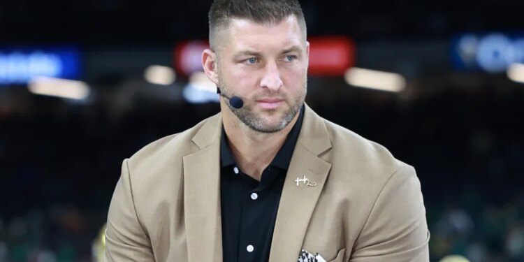 Tim Tebow wearing a tan suit jacket and black shirt with a microphone earpiece.
