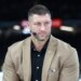 Tim Tebow wearing a tan suit jacket and black shirt with a microphone earpiece.