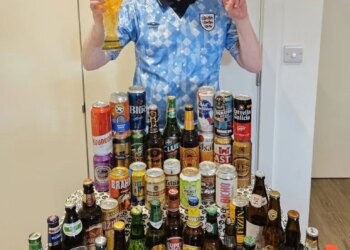 Londoner Gus Hully in a light blue soccer jersey holds up a beer glass and makes a peace sign in front of a table full of various beer bottles and cans from the 48 countries in the World Cup.