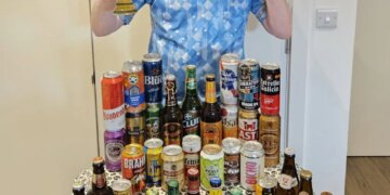 Londoner Gus Hully in a light blue soccer jersey holds up a beer glass and makes a peace sign in front of a table full of various beer bottles and cans from the 48 countries in the World Cup.