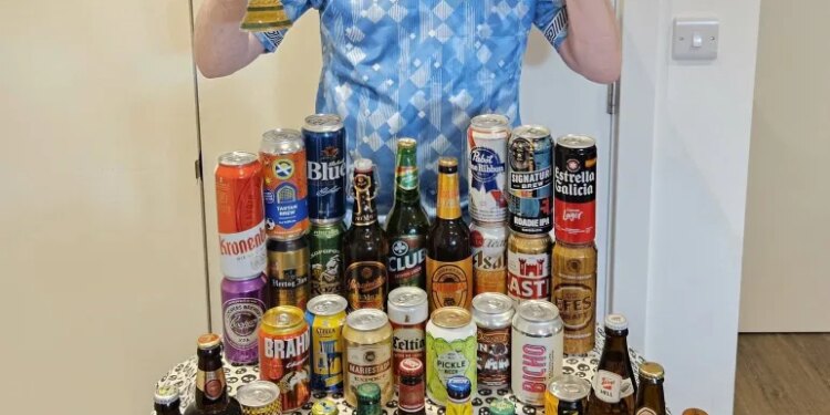 Londoner Gus Hully in a light blue soccer jersey holds up a beer glass and makes a peace sign in front of a table full of various beer bottles and cans from the 48 countries in the World Cup.