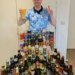 Londoner Gus Hully in a light blue soccer jersey holds up a beer glass and makes a peace sign in front of a table full of various beer bottles and cans from the 48 countries in the World Cup.